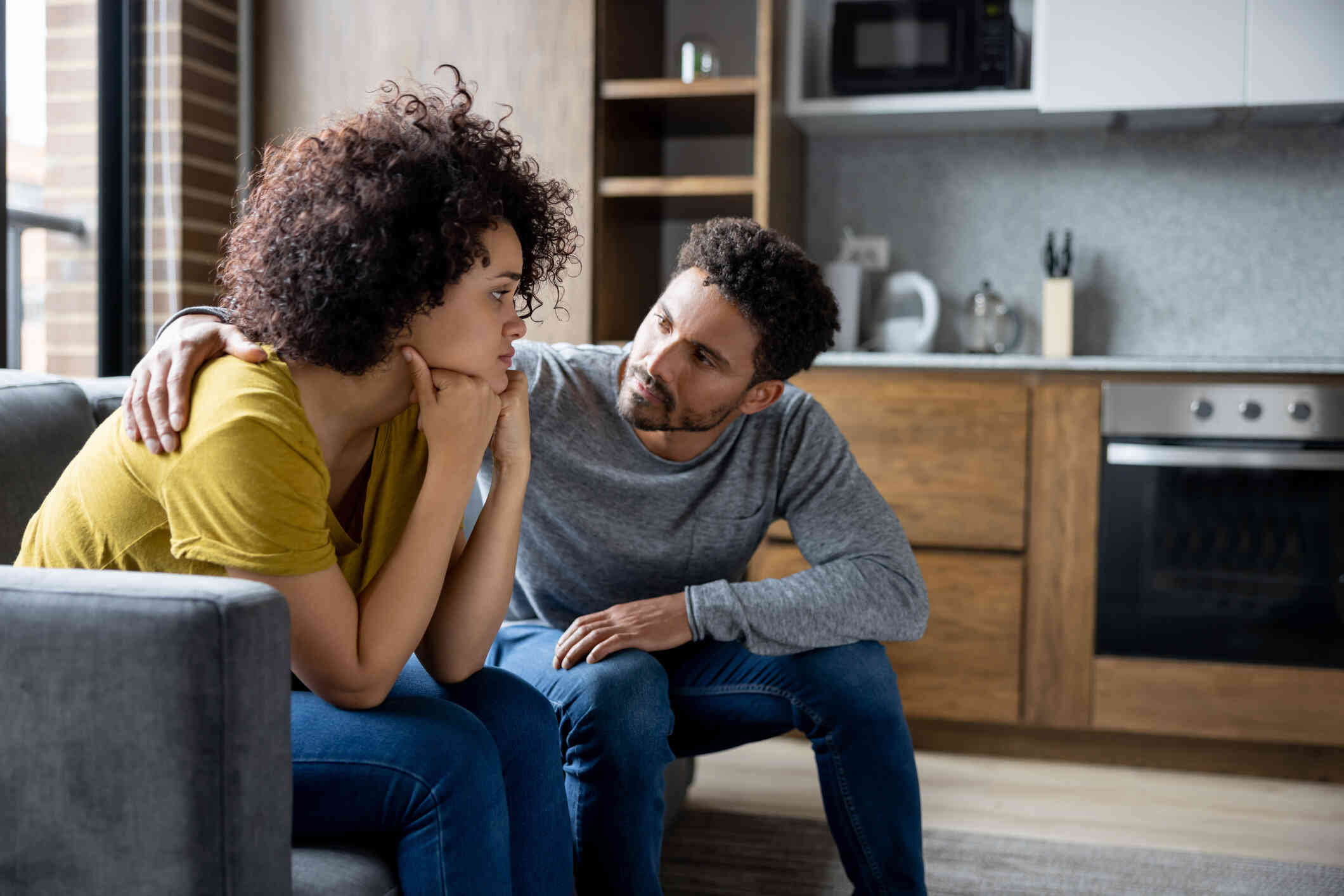 A male and female couple sit next to each other on the couch and have a serious conversation.